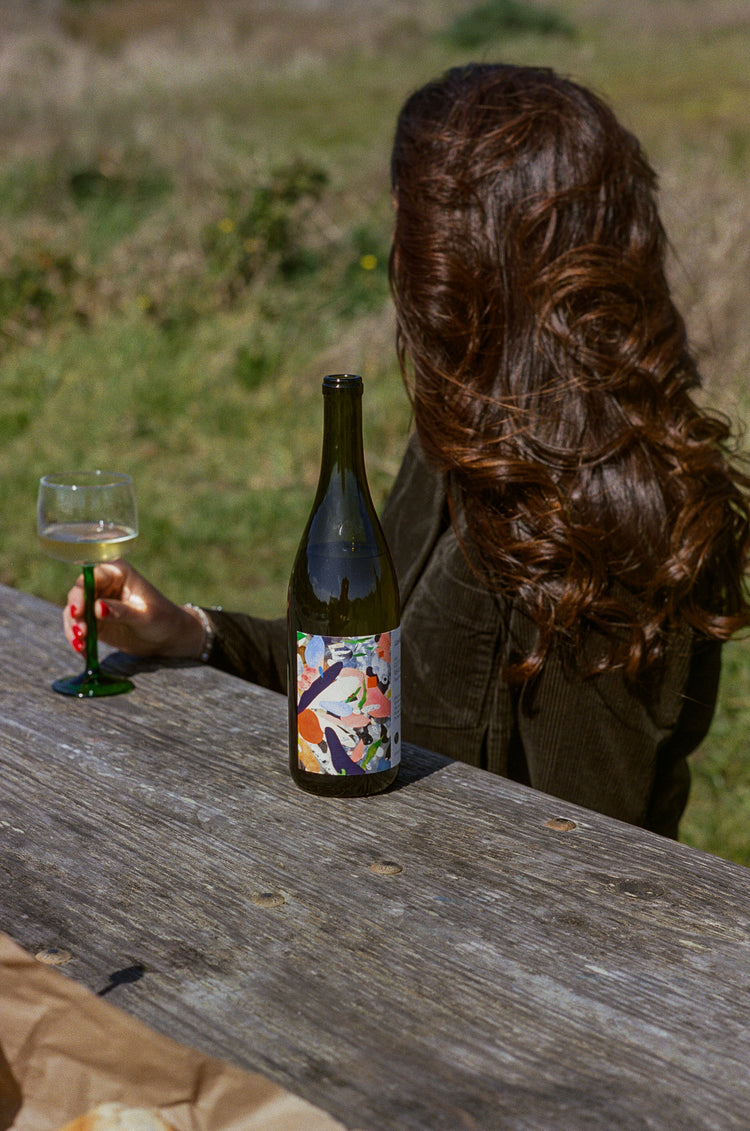 A woman with long hair sits at a picnic table, holding a wine glass. A colorful wine bottle is in front of her, with a grassy field in the background.