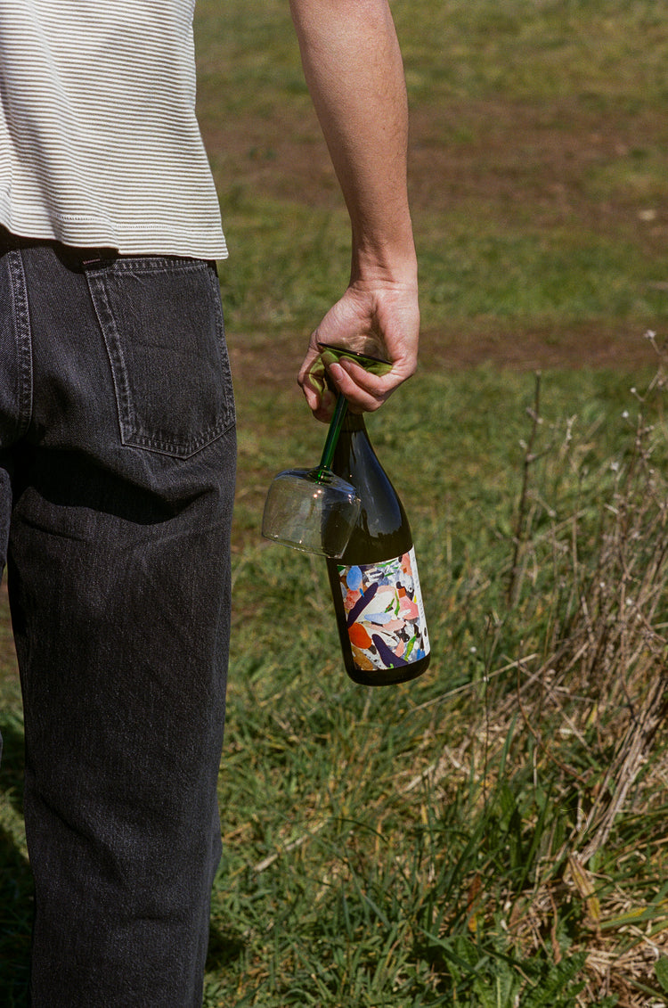 A person stands on grass holding a bottle of Martha Stoumen Out to the Meadow with a colorful label and an upside-down, green stemmed vintage wine glass. Casual outdoor setting, relaxed mood.