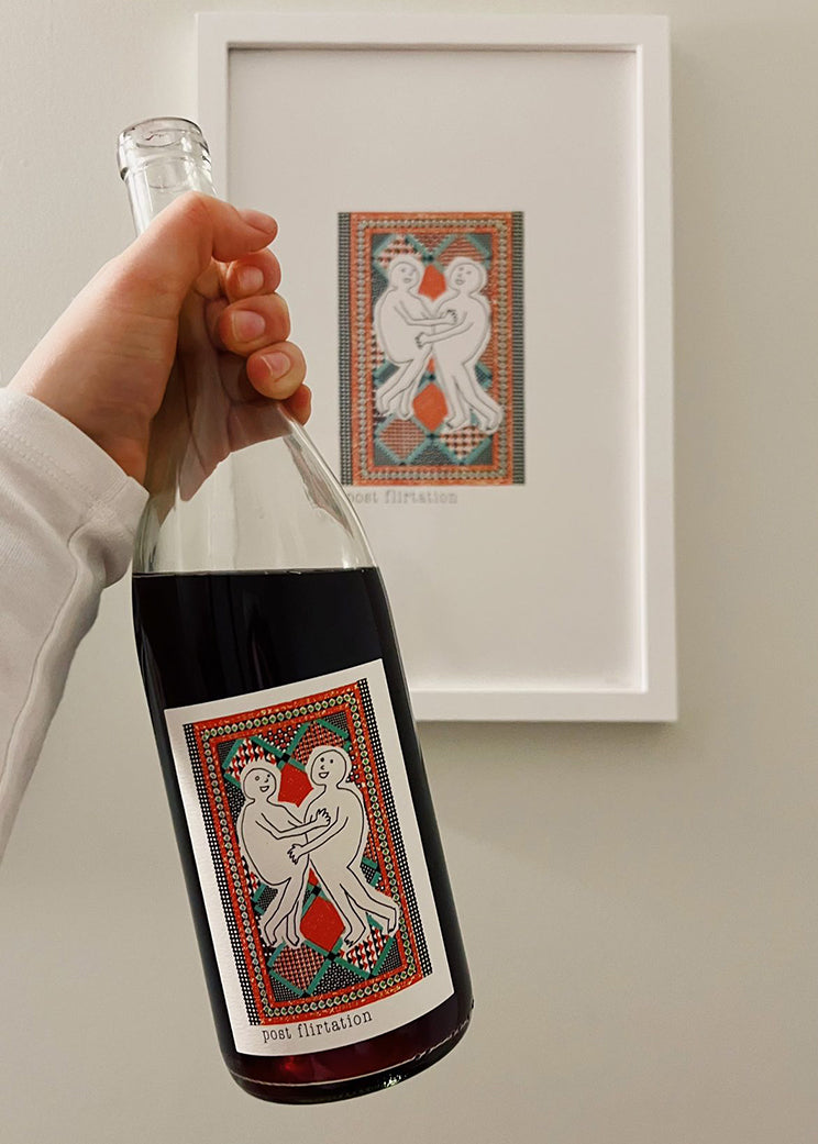 A hand holds a bottle of wine featuring a colorful label with two figures embracing, next to a framed print of the same design.