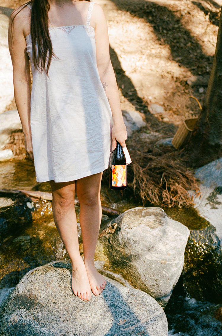 A woman stands on a rock in a creek, wearing a white vintage sun dress, holding a bottle of Martha Stoumen Wines Benchlands 2022 by the neck of the bottle. 